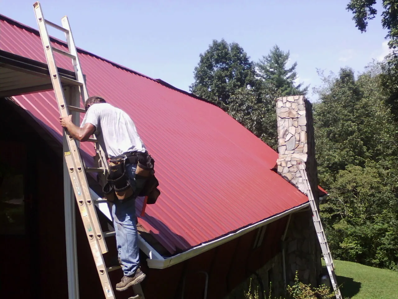 Roofer on a ladder beside a red metal roof and stone chimney — Soffit & Fascia Repair by Roofing Brothers in Corral